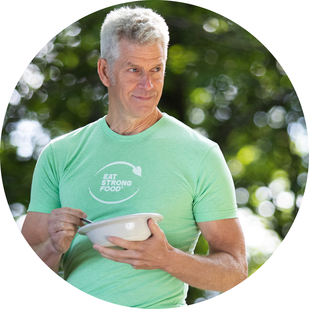 A man with short gray hair, wearing a light green Eat Strong Food t-shirt, smiles while holding a bowl and spoon outdoors against a background of green trees.