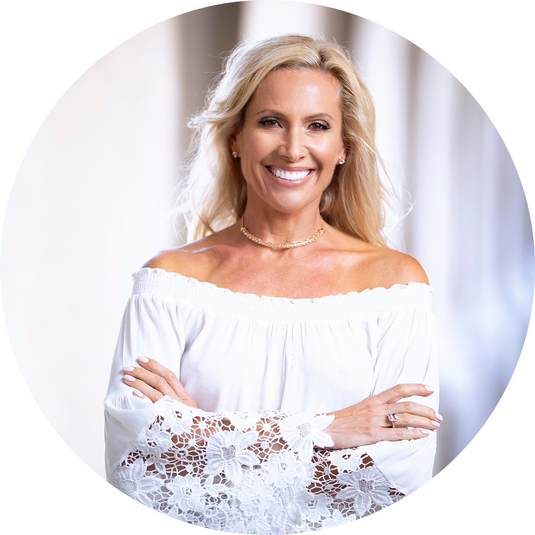 A smiling woman with long blonde hair wears an off-the-shoulder white lace top, standing with arms crossed against a softly blurred light background.