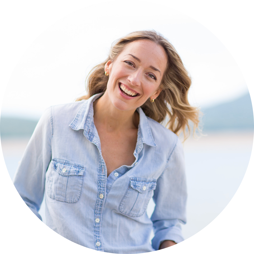 A woman with long, wavy blonde hair wearing a light denim shirt smiles brightly outdoors with a blurred natural background.