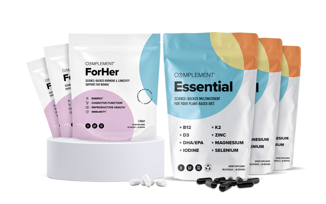 Five pouches of Complement ForHer and four pouches of Complement Essential multivitamins are displayed on a white platform, with white and black capsules scattered in front. The packaging is colorful and modern.