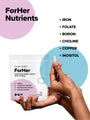 A hand holds a white capsule against a white background beside the ForHer Multinutrient package by Complement, featuring key nutrients like iron, folate, boron, choline, copper, and inositol to support hormonal balance and longevity for women.
