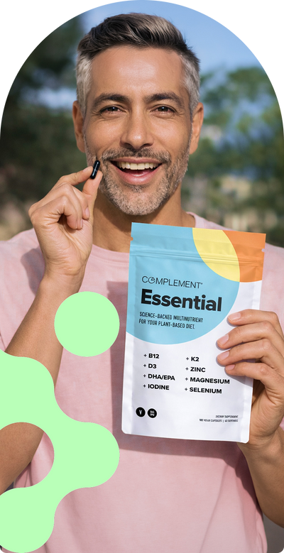 A smiling man wearing a pink shirt holds a small capsule in one hand and a colorful bag of Complement Essential multivitamins in the other, standing outdoors.