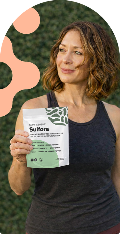 A woman with short brown hair, wearing a dark tank top, stands outdoors holding a Complement Sulfora supplement packet. The background is leafy green with an abstract, peach-colored design on the left.