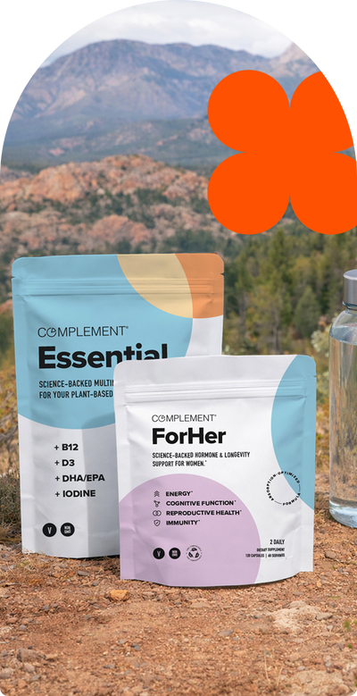 Two supplement pouches labeled “Complement Essential” and “Complement For Her” stand on rocky ground outdoors with mountains and trees in the background. An orange abstract shape is in the sky area.