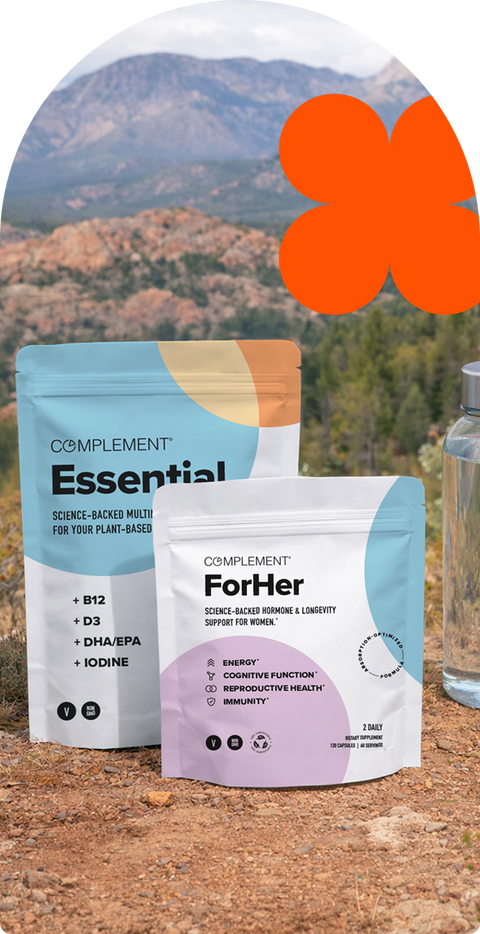 Two supplement pouches labeled “Complement Essential” and “Complement For Her” stand on rocky ground outdoors with mountains and trees in the background. An orange abstract shape is in the sky area.