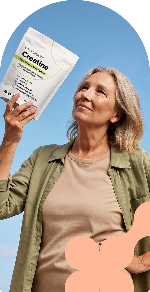 A smiling older woman with blonde hair holds up a white pouch labeled “Complement Creatine” against a blue sky. She wears a tan shirt and olive-green jacket, looking upward with a thoughtful expression.