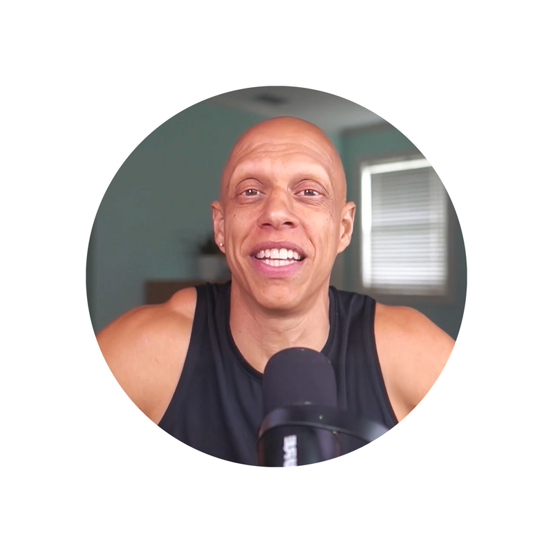 A smiling bald person wearing a black tank top speaks into a microphone, with a room and window in the blurry background. The image is framed in a white circular border.