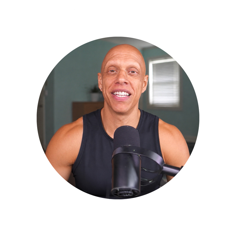 A smiling, bald man in a sleeveless black shirt sits in front of a microphone, speaking, with a softly lit room and window in the background.