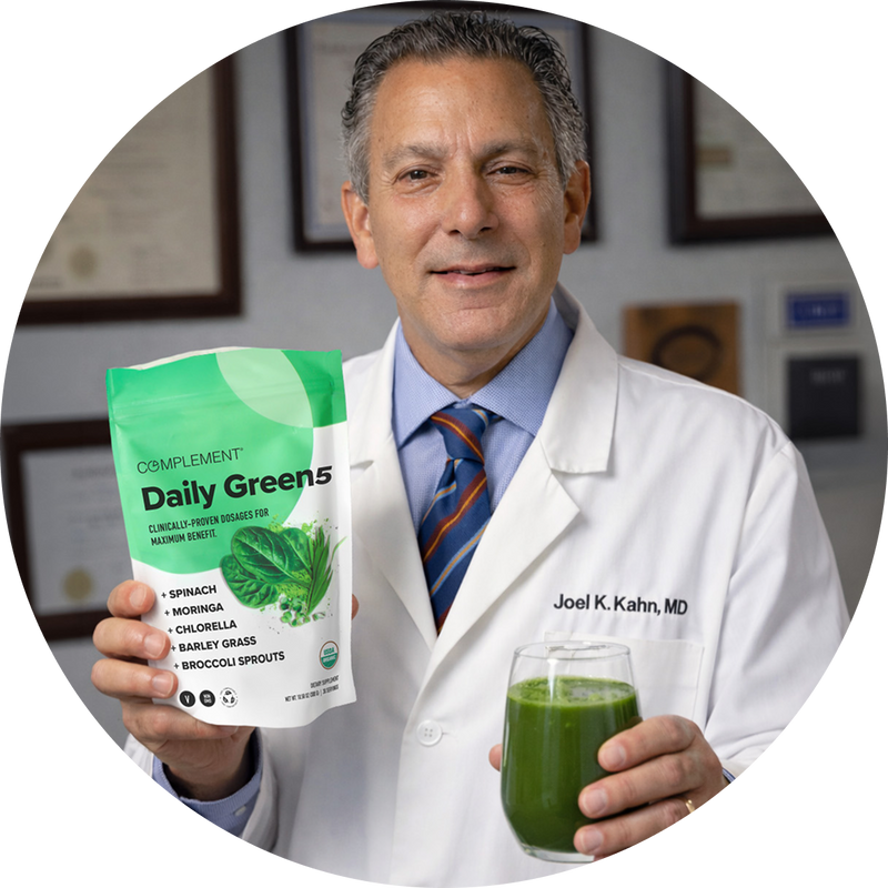 A man in a white lab coat holds a green drink in one hand and a bag labeled “Complement Daily Greens” in the other. The bag lists ingredients like spinach, moringa, and barley grass. Certificates hang on the wall behind him.
