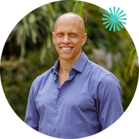 A smiling bald man wearing a light purple button-up shirt stands outdoors in front of green foliage, with a teal starburst graphic in the top right corner.