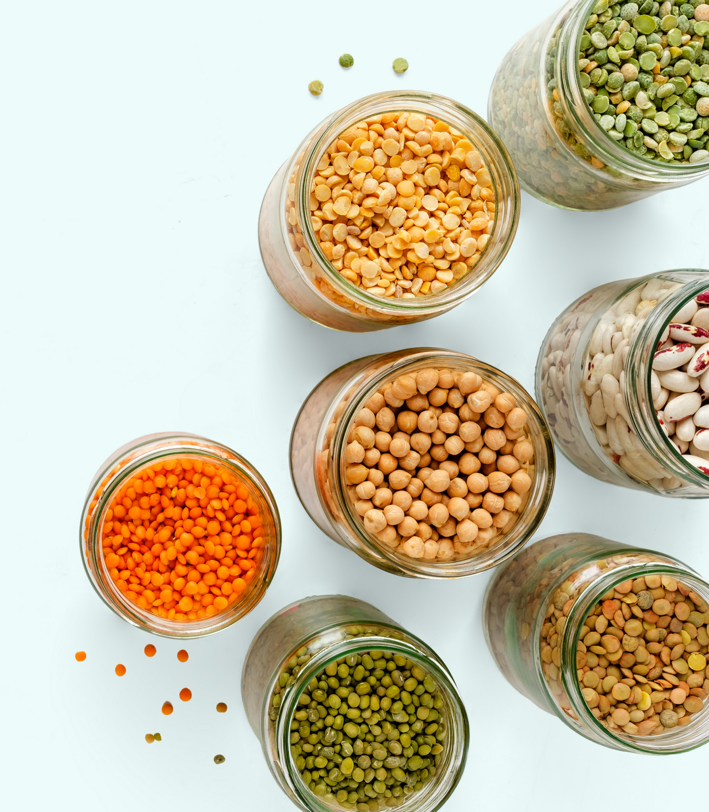 Glass jars filled with various dried legumes, including red, green, and yellow lentils, chickpeas, mung beans, and white beans, arranged on a light background with some lentils scattered nearby.
