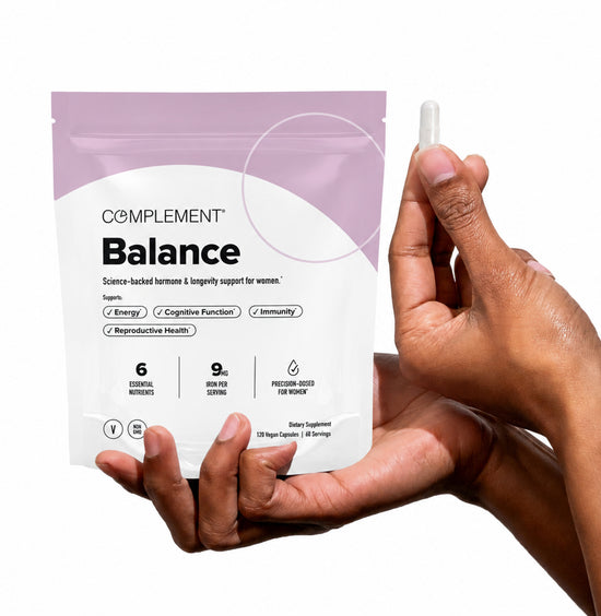 A hand holds a Balance supplement pouch while the other hand holds a capsule. The pouch highlights benefits for womens hormone health, energy, cognitive function, and immunity.
