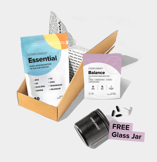 Complement Essential and Balance supplements are displayed with their packaging, inside an open cardboard box. A black glass jar labeled COMPLEMENT is shown nearby with text FREE Glass Jar. Capsules and tablets are scattered.