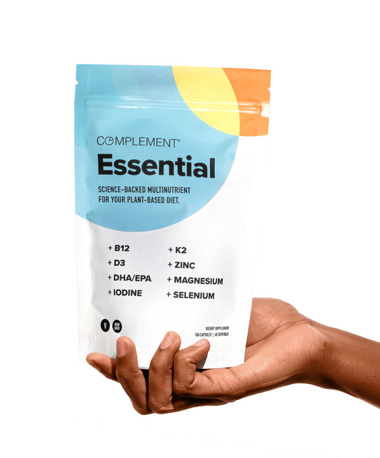 A hand holds a white Complement Essential multivitamin pouch listing nutrients like B12, D3, DHA/EPA, iodine, K2, zinc, magnesium, and selenium. The package has colorful yellow and blue accents.