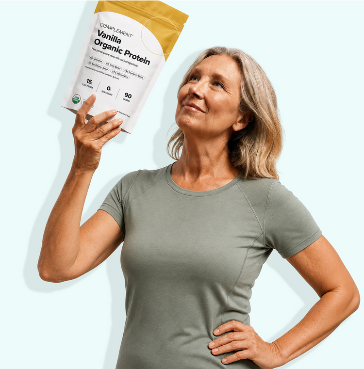 Smiling older woman in a gray shirt holds up a bag of Complement Vanilla Organic Protein against a light background, looking at it with pride.