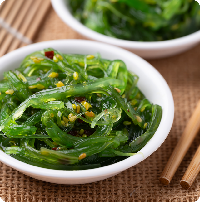 A close-up of a white bowl filled with seasoned seaweed salad, garnished with sesame seeds. In the background, there is a second bowl and a pair of chopsticks resting on a woven placemat.