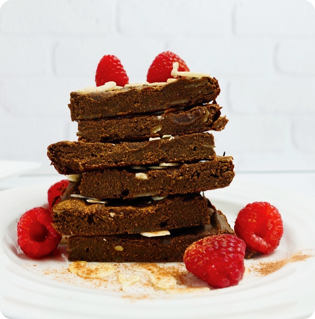 A stack of chocolate brownies topped with almond slivers is on a white plate, surrounded by fresh raspberries and a dusting of cinnamon, against a white background.