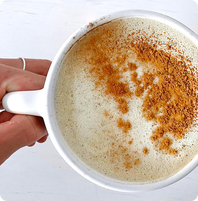 A hand holding a white mug filled with a frothy drink, topped with a sprinkle of cinnamon on one side. The background is a plain white surface.