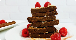 A stack of chocolate brownies garnished with almond slices and raspberries sits on a white plate, with extra raspberries and a dusting of cinnamon around it.