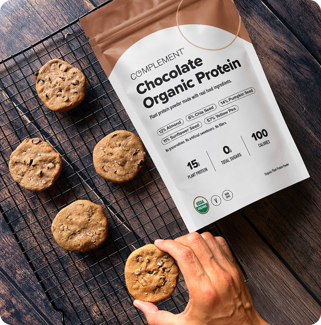A hand grabs a chocolate chip cookie from a cooling rack beside a bag of Complement Chocolate Organic Protein powder on a wooden table.