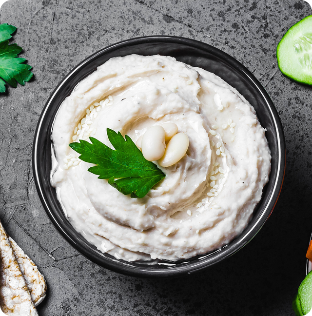 A bowl of creamy white bean dip garnished with parsley, two white beans, and sesame seeds, placed on a dark gray surface with cucumber slices, crackers, and a parsley leaf nearby.