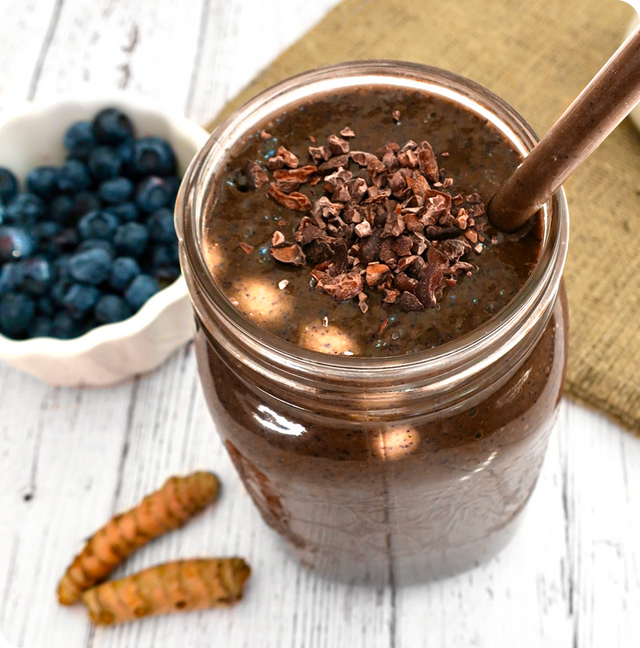 A mason jar filled with a chocolate smoothie, topped with cacao nibs and a straw, sits on a rustic white table near fresh blueberries and turmeric roots.