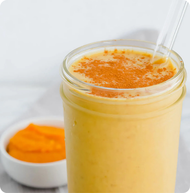 A glass jar filled with a creamy orange smoothie, topped with sprinkled cinnamon, and a clear straw. A small bowl of orange puree is in the background.