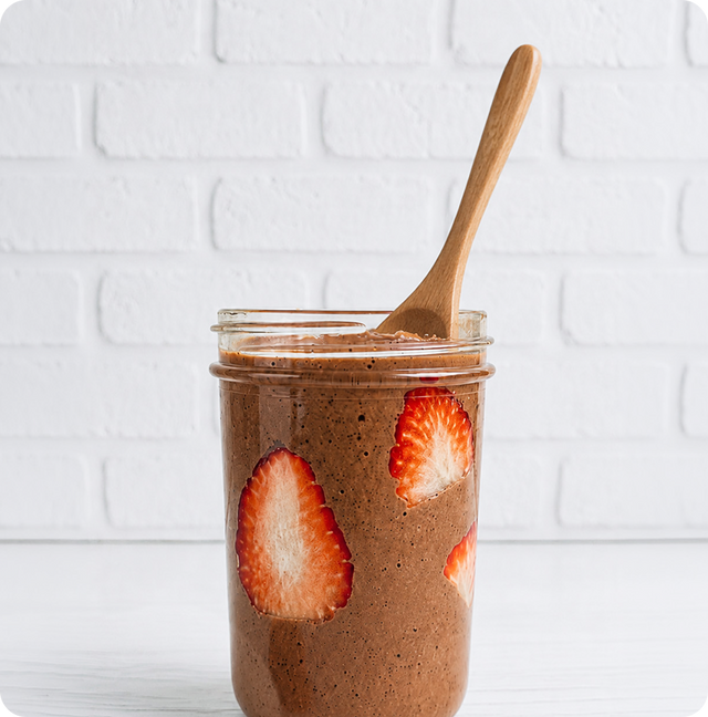 A glass jar filled with chocolate chia pudding and slices of strawberry, with a wooden spoon inside, sits on a white surface against a white brick wall.