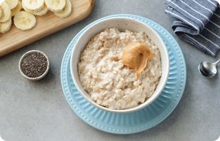 A bowl of oatmeal topped with a scoop of peanut butter, placed on a blue plate. Nearby are sliced bananas on a cutting board, a small bowl of chia seeds, a striped napkin, and a spoon on a light gray surface.