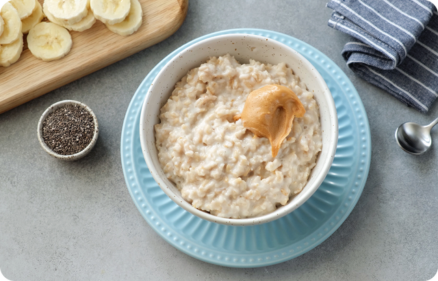 A bowl of oatmeal topped with a scoop of peanut butter, placed on a blue plate. Nearby are sliced bananas on a cutting board, a small bowl of chia seeds, a striped napkin, and a spoon on a light gray surface.