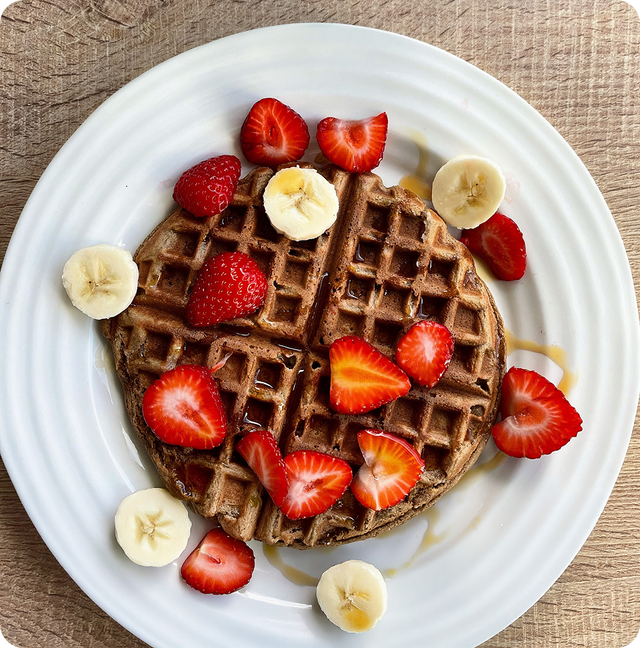 A chocolate waffle topped with sliced strawberries and bananas, drizzled with syrup, served on a white plate on a wooden surface.
