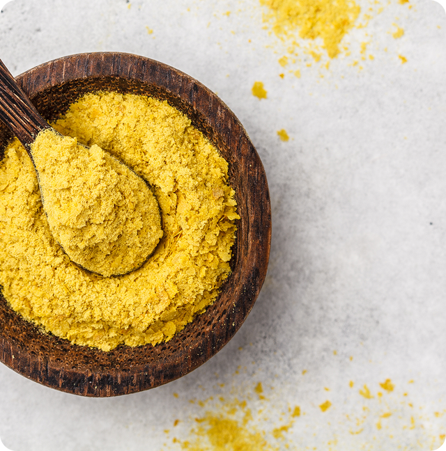 A wooden bowl filled with yellow nutritional yeast flakes, with a wooden spoon resting inside. Some of the yellow flakes are scattered on a light, speckled surface around the bowl.