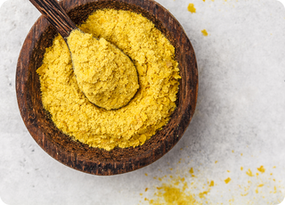 A wooden bowl filled with yellow nutritional yeast flakes, with a wooden spoon resting inside, and some flakes scattered on a light-colored surface.