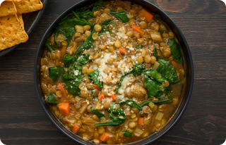 A bowl of hearty lentil soup with spinach, carrots, celery, and onions, topped with grated cheese. Crackers are visible on a plate to the side on a dark wooden table.
