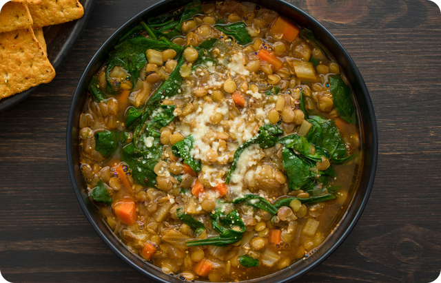A bowl of hearty lentil soup with spinach, carrots, celery, and onions, topped with grated cheese. Crackers are visible on a plate to the side on a dark wooden table.