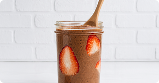 A glass jar filled with chocolate chia pudding, garnished with strawberry slices on the inside, with a wooden spoon resting in the jar. White brick wall background.