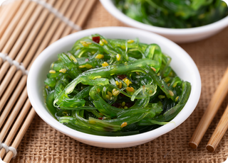 A white bowl filled with bright green seaweed salad garnished with sesame seeds, placed on a woven mat next to chopsticks and a sushi rolling mat. Another bowl of salad is blurred in the background.