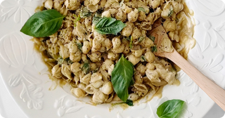 A white plate of shell pasta mixed with pesto sauce, garnished with fresh basil leaves, with a wooden spoon resting on the side.