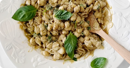 A white plate of shell pasta mixed with pesto sauce, garnished with fresh basil leaves, with a wooden spoon resting on the side.