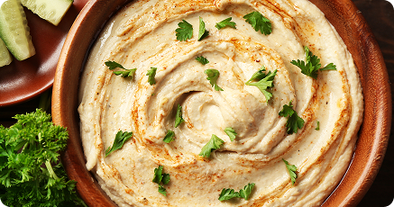 A bowl of creamy hummus garnished with paprika and fresh parsley, with cucumber slices and parsley on the side.