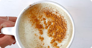 A hand holds a white mug filled with a frothy beverage, topped with a generous sprinkling of cinnamon on one side.