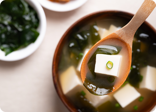 A wooden spoon holds tofu cubes, seaweed, and green onions over a bowl of miso soup. In the background, a small dish contains additional seaweed.