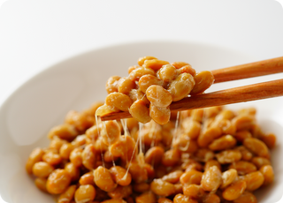 Chopsticks holding up sticky, fermented soybeans (natto) with stringy texture, over a white plate filled with more natto, against a plain white background.