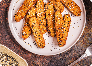 A white plate with several almond cookies sprinkled with white and black sesame seeds, placed on a textured surface. A small bowl of mixed seeds and a fork are nearby.