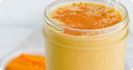 A glass of creamy orange smoothie topped with cinnamon powder, with a clear straw and a small dish of orange fruit puree in the background.