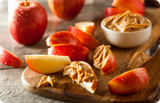 Sliced red apples on a wooden board, some topped with creamy peanut butter, with a small white bowl of peanut butter and whole apples in the background.