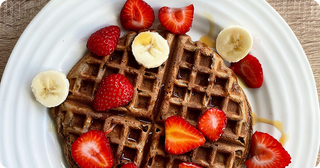 A round waffle topped with sliced strawberries and bananas, drizzled with syrup, served on a white plate.