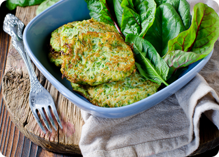 A blue bowl with zucchini fritters and fresh green leafy vegetables, placed on a rustic wooden surface with a beige cloth and a fork beside it.