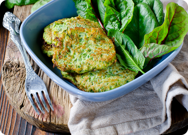 A blue bowl with zucchini fritters and fresh green leafy vegetables, placed on a rustic wooden surface with a beige cloth and a fork beside it.