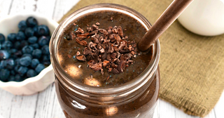 A mason jar filled with a chocolate smoothie, topped with cacao nibs and a metal straw. In the background, fresh blueberries in a white bowl and a glass jar are visible on a rustic wooden surface.